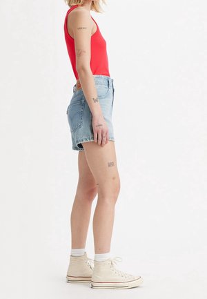 Osoba ubrana w czerwony tank top, jasnoniebieskie dżinsowe szorty, białe skarpetki i kremowe sneakersy za kostkę, z widocznymi tatuażami na ramieniu i nodze.