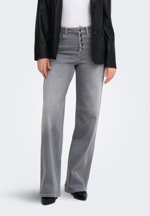 Kvinde iført gråvidde jeans med høj talje og fem guldknapper, kombineret med en hvid top, en sort blazer og sorte spidse sko.
