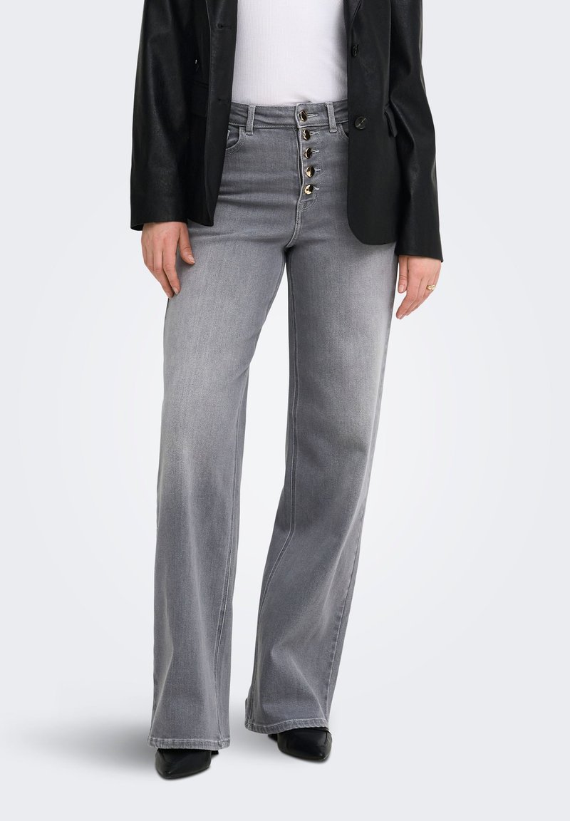 Femme portant un jean gris taille haute à jambes larges avec cinq boutons dorés, associé à un top blanc, un blazer noir et des chaussures noires pointues.