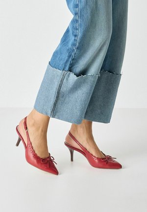 Czerwone skórzane sandały na obcasie z paskiem z tyłu i zaostrzonym noskiem, zdobione kokardką i wycięciami. Noszone z jeansami o postrzępionych zakończeniach.