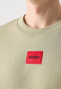 Gros plan sur un sweat-shirt vert clair à col rond avec un patch carré rouge portant le texte noir "HUGO" sur la poitrine.