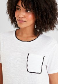 Woman with curly hair wearing a white t-shirt featuring black zigzag trim on the neckline and front pocket, looking slightly right.