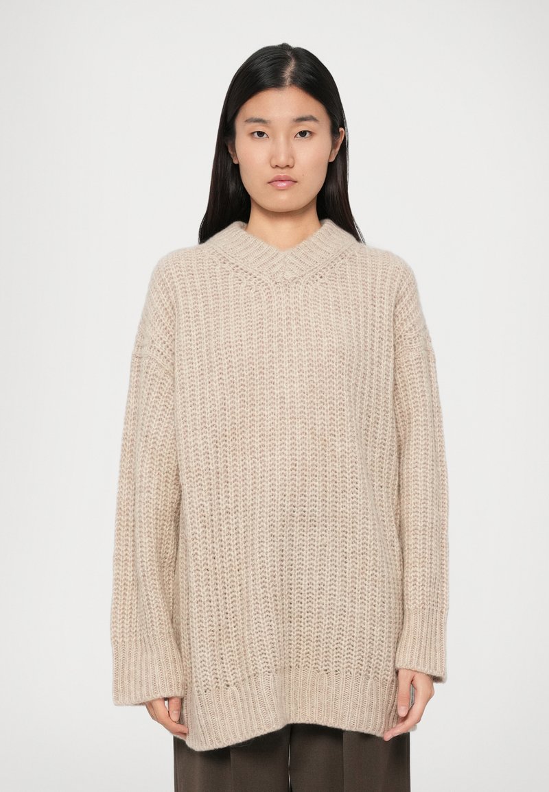 Beige gestrickter Pullover mit lockerer Passform, V-Ausschnitt, gerippter Textur und fallenden Schultern. Lange Ärmel reichen bis zu den Handgelenken.