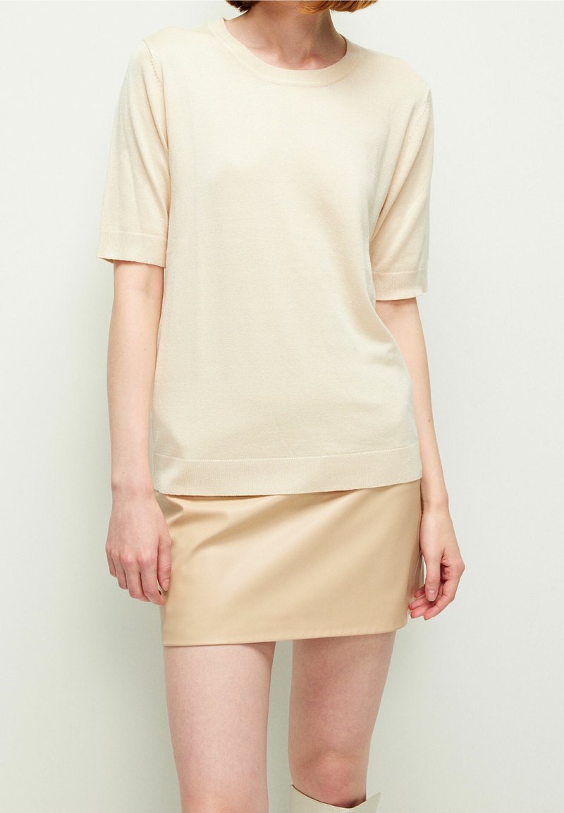 Femme portant un haut en maille beige à manches courtes et une minijupe en cuir beige clair, debout devant un fond uni clair.