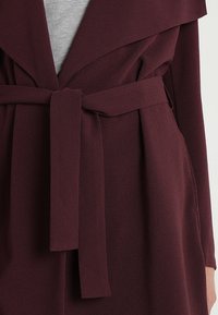 Burgundowa kurtka z szerokim kołnierzem, paskiem w talii oraz miękkim, teksturowanym materiałem. Posiada kieszenie i luźny krój.