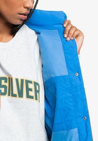 Blue lightweight jacket with contrasting fabric panels, snap button detail, and a smooth texture, worn over a grey sweatshirt with bold text.