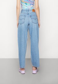Jeans de tiro alto en azul claro con piernas anchas, dos bolsillos traseros y un parche de cuero marrón en la cinturilla. Lleva zapatos de plataforma lavanda.