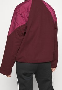 Maroon jacket with a shiny purple upper section, featuring a loose fit, long sleeves, and a rounded back hem. Textured fabric.