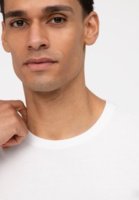 Weißes Rundhals-T-Shirt, glattes Material, eng anliegend am Hals, mit kurzen Ärmeln; die Hand des Modells berührt den Halsauschnitt.