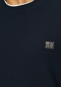 Marineblauer, strukturierten Pullover mit einem gerippten Rundhalsausschnitt und einem schwarzen quadratischen Logopatch mit der Aufschrift "s.O." auf der linken Seite.