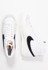 Baskets montantes avec un dessus en cuir blanc, logo swoosh noir, accents en daim gris et semelle en caoutchouc blanc. Les lacets sont blancs.