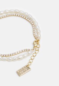 Guldtonad armband med två strängar: en med pärlivriga pärlor och en annan med klara kristalldetaljer, avslutad med ett märke och lås.