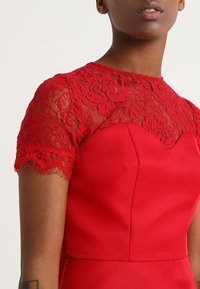 Röd klänning med en figursydd överdel och korta ärmar i blommig spets. Slät tyg med en strukturerad yta och intrikata spetsdetaljer vid axlarna.