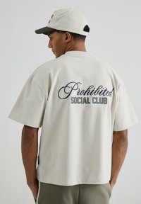 Cămașă oversized bej deschis cu mâneci scurte, având inscripția "Prohibited Social Club" în stil cursiv de culoare albastru închis pe spate. Material din bumbac.