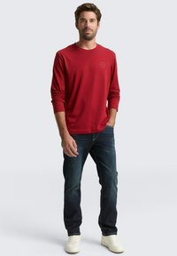 Rode longsleeve shirt met ronde halslijn en logo op de borst, gecombineerd met donkere jeans en witte sneakers, staand tegen een neutrale achtergrond.
