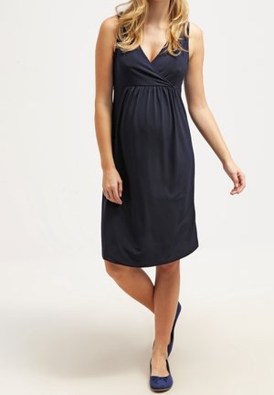 Frau trägt ein ärmelloses marineblaues knielanges Kleid mit V-Ausschnitt und marineblaue Ballerinas, steht mit einem Bein vor dem anderen überkreuzt.