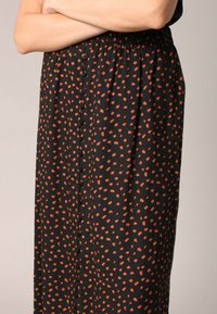 Femme portant une jupe noire avec des points orange irréguliers et un détail de boutons sur le devant, les bras croisés sur la taille.