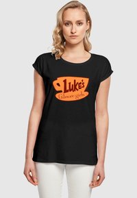 Must черн футболка lühikeste varrukatega, millel on oranži kohvikruusi graafika ja julge tekst, mis ütleb "Luke'i Gilmore tüdrukud."