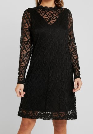 Kvinde iført en knælængde sort blondekjole med lange, gennemsigtige ærmer og et blomsterprint, stående mod en ensfarvet baggrund.