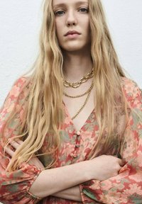 Giovane donna con lunghi capelli biondi che indossa catene d'oro a strati e una camicetta floreale color corallo con bottoni, con le braccia incrociate su uno sfondo bianco semplice.