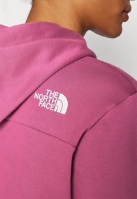 Hanorac roz cu glugă și o textură fină. Are logo-ul "The North Face" în alb pe umăr, evidențiind identitatea mărcii.