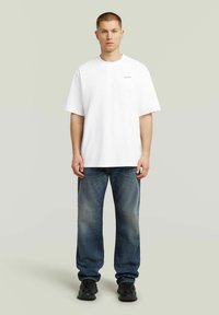 T-shirt bianca in cotone con scollo rotondo e maniche corte, abbinata a jeans blu chiaro a gamba dritta e sneakers nere.