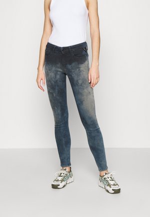 Jeans ajustados de color azul desgastado con una textura rasgada, que cuentan con bolsillos traseros y una cremallera. Combinados con zapatillas deportivas blancas.