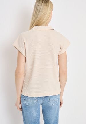 Femme blonde portant un haut texturé beige clair à manches courtes avec col, associé à un jean en denim bleu clair, vue de dos.