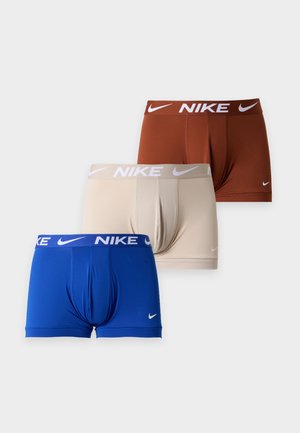 Veltrænet mand stående barfodet iført beige Nike boxer briefs mod en ensfarvet lys grå baggrund.