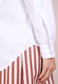 Vit bomullsskjorta med rundad nederkant och knäppta ärmar, ihopparad med en plisserad mörkröd och vit randig kjol. Synlig hand som greppar tyget.