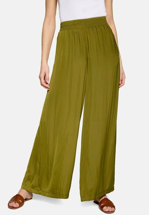 Femme portant un pantalon large vert olive associé à un haut blanc et des sandales marron sans bride, debout devant un fond uni.