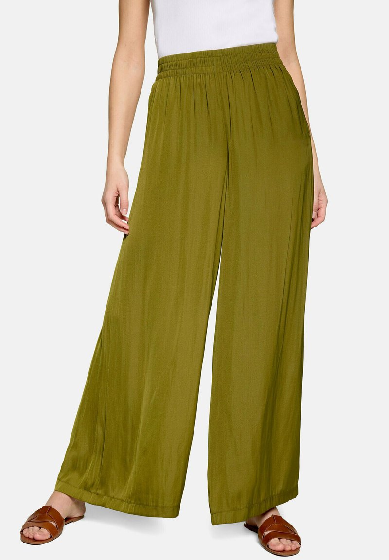 Femme portant un pantalon large vert olive associé à un haut blanc et des sandales marron sans bride, debout devant un fond uni.