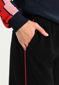 Svart träningsbyxor med slim fit, som har ett rött sidofält, och en hand i fickan på högra sidan. Standard midjeband.