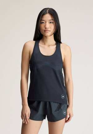 Young woman with long dark hair wearing black athletic tank top and shorts standing against plain light background.