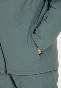 Herrars grön vattentät jacka med texturerad tyg, blixtlås och sidofickor. Har en figursydd design med justerbara ärmslut och nederkant.