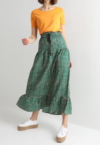 Jupe maxi verte à motifs avec un ourlet à volants et une taille élastique, associée à un haut orange à manches courtes et des baskets blanches.