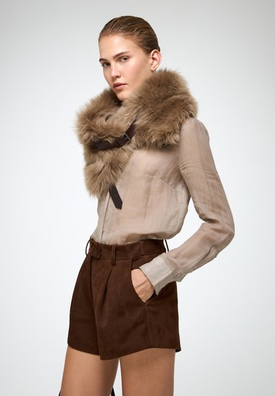Donna che indossa una blusa beige trasparente, pantaloni corti in suede marrone e un ampio collare di pelliccia marrone con fibbia, in posa con una mano in tasca.