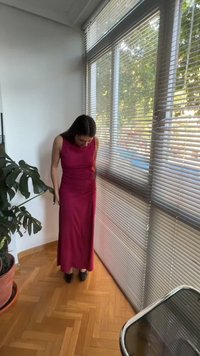 Femme en longue robe rouge sans manches debout à l'intérieur près de grandes fenêtres avec des stores et une plante en pot sur un sol en bois.
