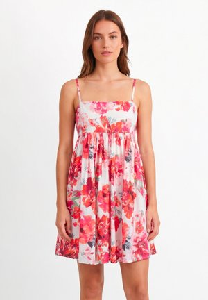 Kobieta w białej sukience bez rękawów do kolan z jasnoczerwonym i różowym kwiatowym wzorem, stojąca na białym tle.