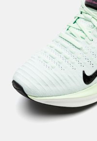 Gros plan d’une chaussure de sport en maille vert clair avec logo Nike noir et lacets, montrant la tige texturée et la semelle intermédiaire blanche.