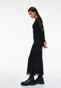 Frau in einem langen schwarzen Kleid aus floralem Spitze mit durchsichtigen Ärmeln und schwarzen klobigen Plateaustiefeln, stehend vor einem schlichten weißen Hintergrund.
