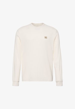 Chemise à manches longues couleur crème clair en tissu texturé, avec un col rond, des poignets côtelés et un petit logo carré sur la poitrine.