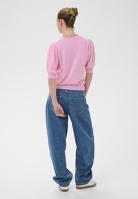 Žena s vlasy staženými do drdolu, oblečená v růžovém tričku s nafouklými rukávy, volných modrých džínách a bílých teniskách, stojící zády k pozadí.