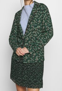 Blazer verde a motivo e gonna coordinata con un design floreale, abbinati a una blusa di un blu chiaro strutturato sotto. Ogni pezzo presenta dettagli intricati.