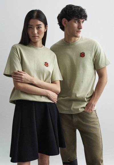 T-shirt color khaki con patch rosso ricamato, indossate da due modelli. Un modello in una gonna nera testurizzata; l'altro in pantaloni beige.