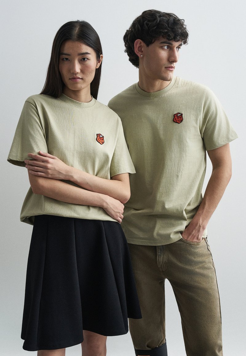 Καφέ T-shirts με κεντημένο κόκκινο λογότυπο, φορεμέν α από δύο μοντέλα. Ένα μοντέλο σε μαύρη υφασμάτινη φούστα; το άλλο σε μπεζ παντελόνι.