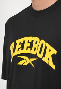 Melnas kokvilnas T-krekls ar lielu dzeltenu "REEBOK" logotipu un grafisko dizainu. Arapa kakls, īsas piedurknes, gluda tekstūra.