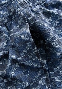 Modrá vzorovaná tkanina s textúrovaným povrchom, s kockovaným vzorom v tmavších a svetlejších odtieňoch, zobrazujúca detail elastického pásu.