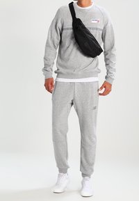 Felpa grigia con scollo a girocollo e striscia orizzontale, abbinata a pantaloni della tuta grigi coordinati. Borsa a tracolla nera indossata sulla spalla. Sneakers bianche.