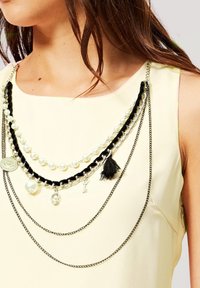 Collar de capas negro y plateado con perlas, cadenas y charms, llevado sobre una blusa amarilla clara. Incluye un borlas y varios acentos metálicos.